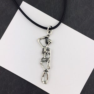 May include: A silver-toned metal pendant shaped like a human skeleton in a standing pose, suspended from a thin black cord. The skeleton figure has articulated limbs and a detailed rib cage, with a small metal loop at the top for the cord attachment.