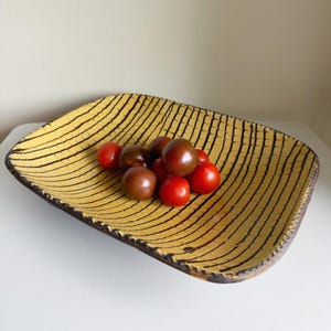 May include: A rectangular, handmade ceramic bowl with a yellow interior and black linear pattern. The bowl contains a mix of red and dark brown cherry tomatoes. The rim of the bowl is dark brown.