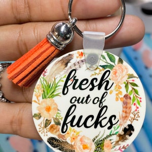 May include: A round keychain with the text "fresh out of fucks" in black script, surrounded by a floral and feather design. It has a silver keyring and an orange tassel.