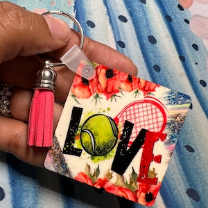 Puede incluir: Un llavero de borla rosa con un dije cuadrado que presenta una raqueta de tenis, una pelota de tenis y la palabra "LOVE" en letras negras. El dije tiene un fondo de acuarela con flores rojas.