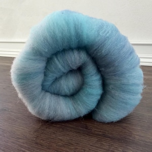 May include: A tightly coiled bundle of blue and teal wool roving. The wool is a soft, fluffy texture, with a gradient of colors from light to dark. The roving is sitting on a dark wood surface.
