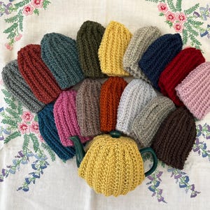 May include: A selection of ribbed knit beanies in a variety of colours, such as red, green, yellow, and blue, are arranged around a yellow knitted tea cosy with a green handle. The items are displayed on a white tablecloth with floral embroidery.