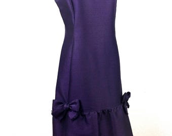 May include: A sleeveless, knee-length purple dress with a round neckline. The dress features a ruffled hemline and two decorative bows. The dress is displayed on a mannequin.