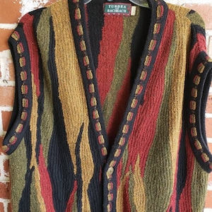Può includere: Un maglione senza maniche con un design colorato e strutturato. Il maglione presenta strisce verticali in nero, rosso, verde oliva e oro. Il colletto e i bordi sono rifiniti con un colore scuro e cuciture decorative. L'etichetta recita "Tundra Bachrach."