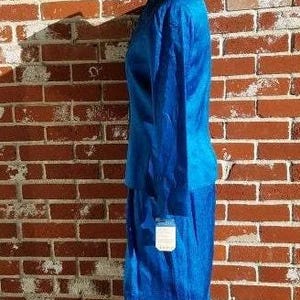 May include: A vibrant blue two-piece suit, consisting of a long-sleeved jacket and a knee-length skirt. The suit is displayed on a mannequin against a brick wall. A tag hangs from the skirt. The fabric has a subtle texture.