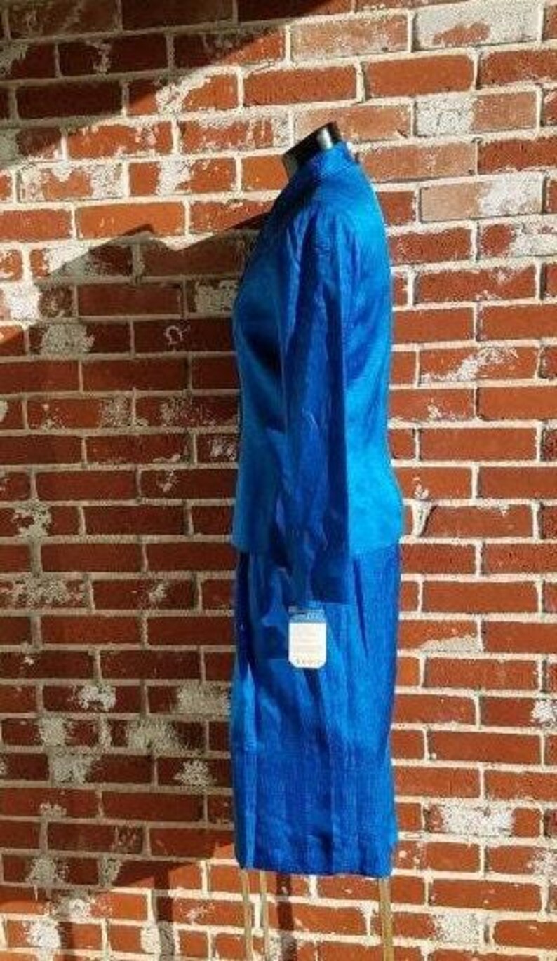 May include: A vibrant blue two-piece suit, consisting of a long-sleeved jacket and a knee-length skirt. The suit is displayed on a mannequin against a brick wall. A tag hangs from the skirt. The fabric has a subtle texture.
