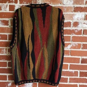 Puede incluir: Un su&eacute;ter sin mangas con un patr&oacute;n abstracto y colorido. El dise&ntilde;o presenta rayas verticales en tonos de rojo, negro, verde oliva y beige. El su&eacute;ter tiene un ribete negro alrededor del cuello, las sisas y el dobladillo. La prenda est&aacute; colgada contra una pared de ladrillos.