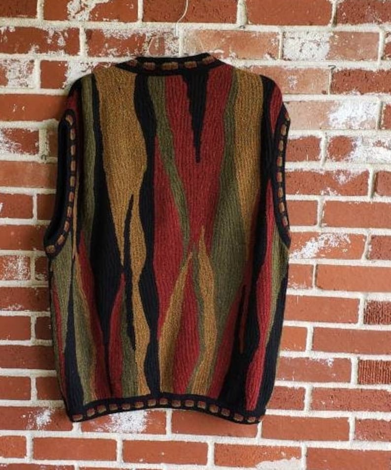 Puede incluir: Un su&eacute;ter sin mangas con un patr&oacute;n abstracto y colorido. El dise&ntilde;o presenta rayas verticales en tonos de rojo, negro, verde oliva y beige. El su&eacute;ter tiene un ribete negro alrededor del cuello, las sisas y el dobladillo. La prenda est&aacute; colgada contra una pared de ladrillos.