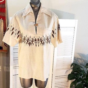 May include: A beige short-sleeved shirt with a brown patterned design of stylised figures. The shirt has a collar and a button closure at the neck.