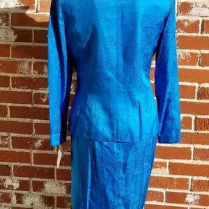 May include: A vibrant blue two-piece suit, featuring a long-sleeved jacket with a mandarin collar and a matching knee-length skirt. The suit has a textured fabric and is displayed on a mannequin against a brick wall.