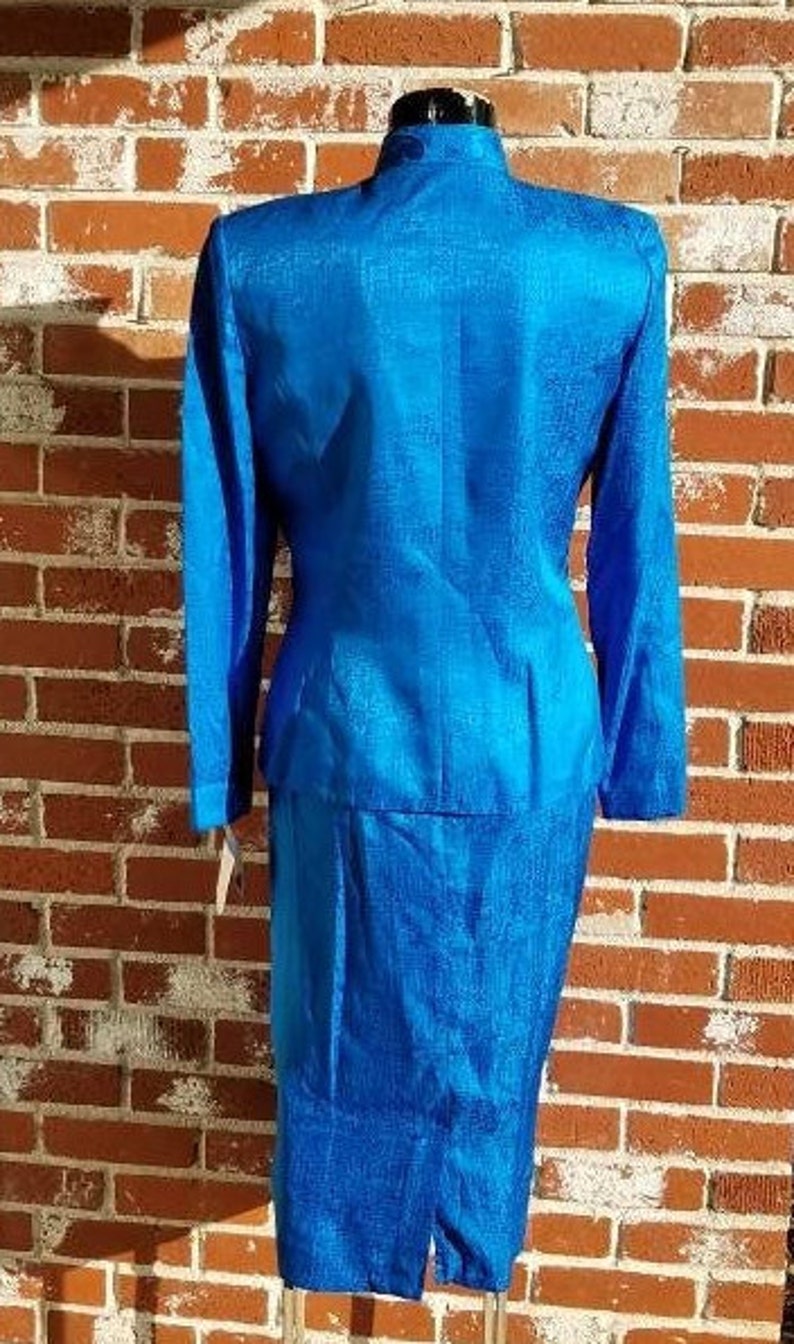 May include: A vibrant blue two-piece suit, featuring a long-sleeved jacket with a mandarin collar and a matching knee-length skirt. The suit has a textured fabric and is displayed on a mannequin against a brick wall.