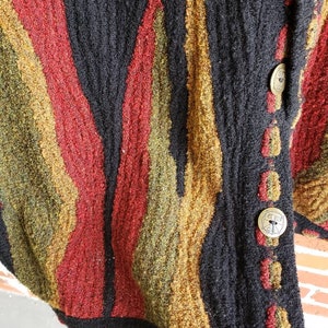 Puede incluir: Primer plano de un c&aacute;rdigan negro texturizado con rayas verticales en rojo, dorado y verde oliva. El c&aacute;rdigan tiene botones dorados grandes y redondos con un dise&ntilde;o decorativo. La tela parece ser un tejido grueso.
