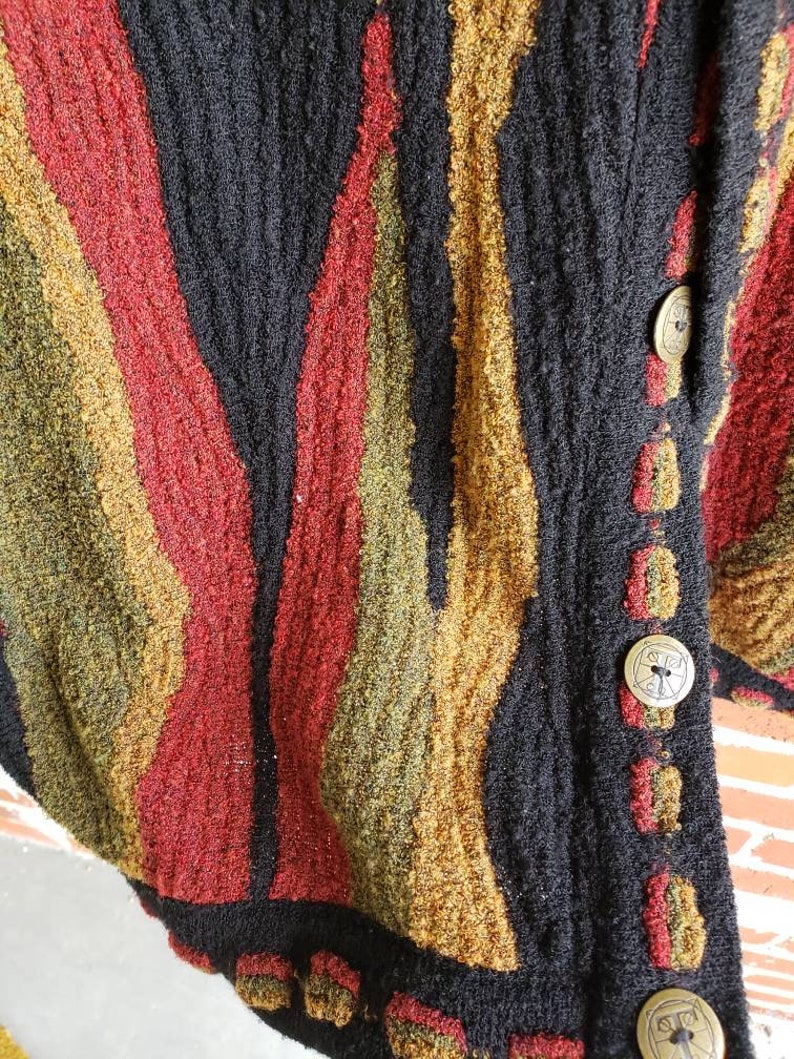Puede incluir: Primer plano de un c&aacute;rdigan negro texturizado con rayas verticales en rojo, dorado y verde oliva. El c&aacute;rdigan tiene botones dorados grandes y redondos con un dise&ntilde;o decorativo. La tela parece ser un tejido grueso.