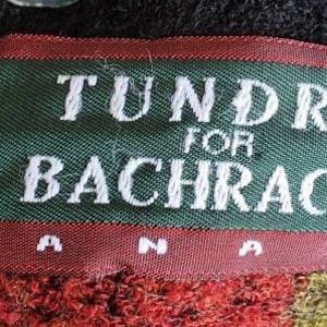 Puede incluir: Una etiqueta rectangular con el texto "TUNDRA FOR BACHRACH CANADA" en letras blancas sobre un fondo verde, enmarcada por un borde rojo. La etiqueta est&aacute; cosida sobre una tela texturizada.