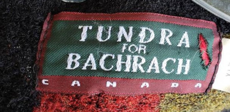 Puede incluir: Una etiqueta rectangular con el texto "TUNDRA FOR BACHRACH CANADA" en letras blancas sobre un fondo verde, enmarcada por un borde rojo. La etiqueta est&aacute; cosida sobre una tela texturizada.