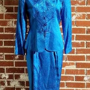 May include: A vibrant blue two-piece suit, featuring a long-sleeved jacket with ornate embroidery and a matching knee-length skirt. The jacket has a mandarin collar and button closure. The suit is displayed on a mannequin against a brick wall.