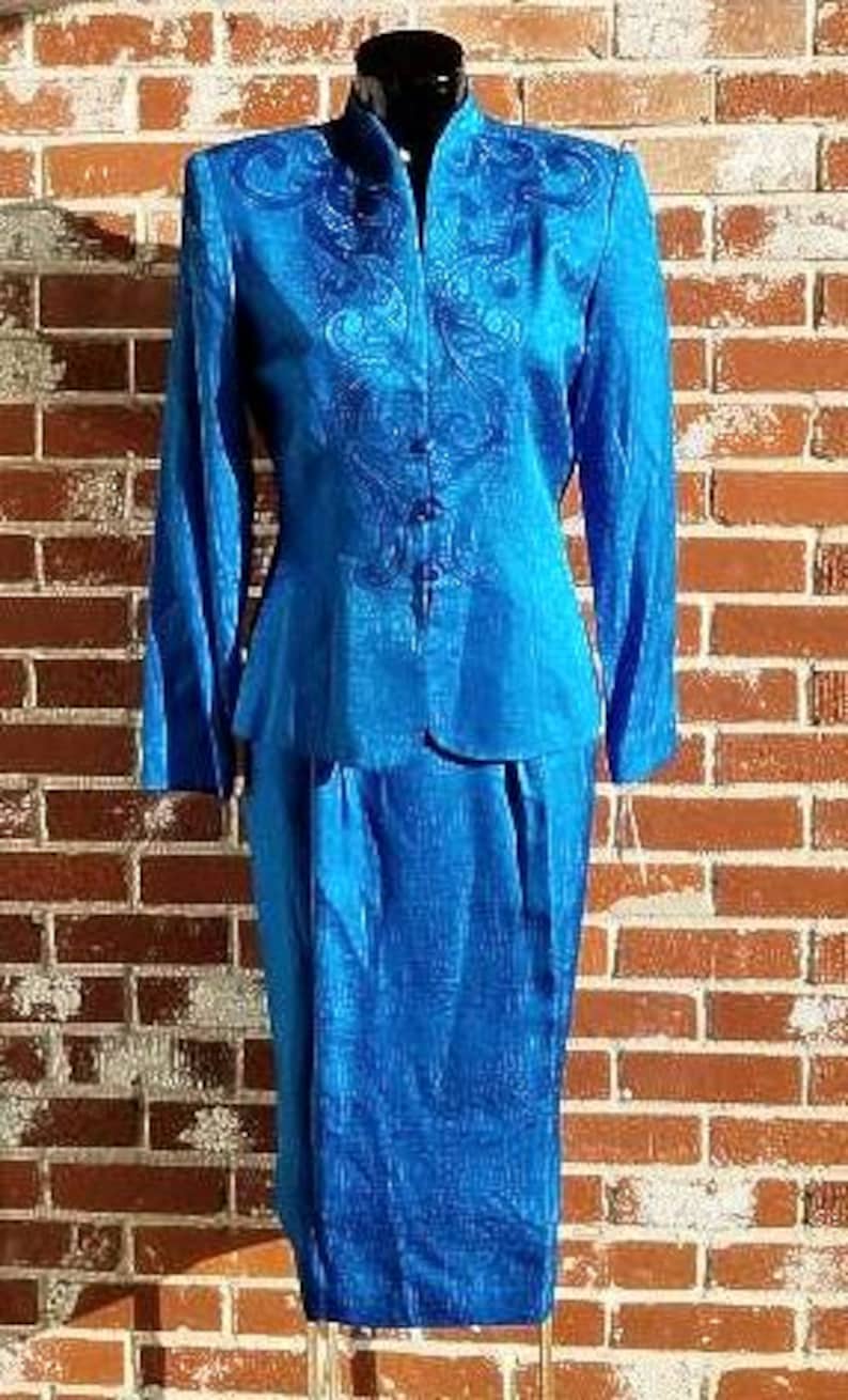 May include: A vibrant blue two-piece suit, featuring a long-sleeved jacket with ornate embroidery and a matching knee-length skirt. The jacket has a mandarin collar and button closure. The suit is displayed on a mannequin against a brick wall.
