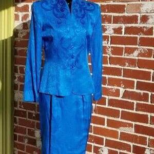 May include: A vibrant blue two-piece suit, consisting of a long-sleeved jacket with ornate embroidery and a matching knee-length skirt. The suit is displayed on a mannequin against a brick wall.