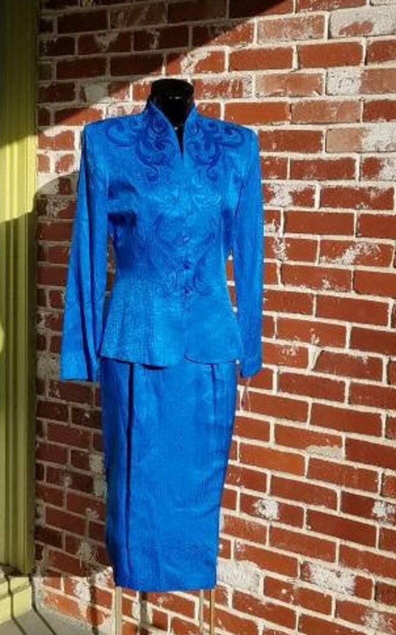 May include: A vibrant blue two-piece suit, consisting of a long-sleeved jacket with ornate embroidery and a matching knee-length skirt. The suit is displayed on a mannequin against a brick wall.