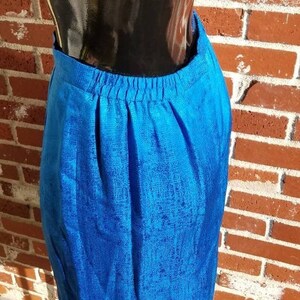 May include: A vibrant blue skirt with a textured pattern. The skirt has an elastic waistband and a knee-length hemline. The fabric appears to have a subtle sheen, catching the light. The skirt is displayed against a brick wall.
