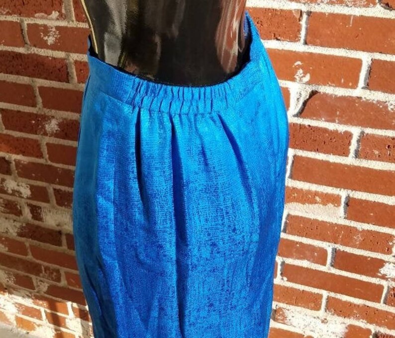 May include: A vibrant blue skirt with a textured pattern. The skirt has an elastic waistband and a knee-length hemline. The fabric appears to have a subtle sheen, catching the light. The skirt is displayed against a brick wall.