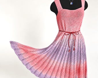 Puede incluir: Un vestido de ganchillo sin mangas con un patrón de color degradado que va del rosa coral en la parte superior al morado claro en el dobladillo. El vestido tiene escote cuadrado, falda acampanada y un cordón rojo oscuro en la cintura.