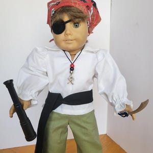 May include: A doll dressed as a pirate, wearing a red bandana with a black eye patch, white shirt, green pants, and a black sash. The doll holds a black telescope and a wooden sword. A necklace with a skull and crossbones charm is also visible.