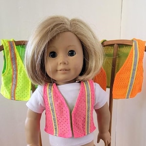 May include: A doll wearing a pink safety vest with reflective stripes. Two additional safety vests in green and orange are hanging in the background.
