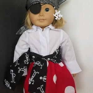 May include: A doll wearing a white shirt, a red and white polka dot skirt, a black and white skeleton print scarf, a black eye patch, and a black and silver bandana with a white skull and crossbones earring.