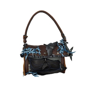 May include: A brown leather handbag with a top handle. The bag features a flap closure and is adorned with dark blue fabric accents, silver studs, and decorative butterflies. The bag has a dark brown handle.