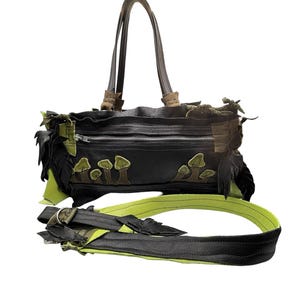 May include: Black leather handbag with a zip closure, featuring green mushroom appliqués and accents. The bag has two brown handles and a detachable shoulder strap with green and black details. The bag is designed with a unique, nature-inspired aesthetic.