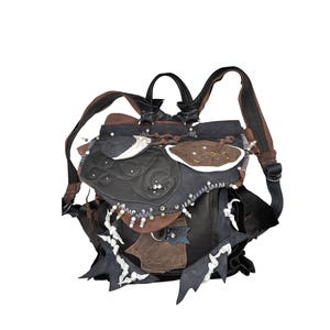 May include: A unique, handcrafted backpack featuring a patchwork design in shades of black, brown, and white. The bag has adjustable straps, a top handle, and decorative embellishments including beads and stitched details. The overall design is artistic and unconventional.