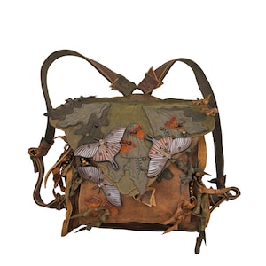 Könnte beinhalten: Handgefertigter Lederrucksack mit einem von der Natur inspirierten Design. Der Rucksack hat eine Mischung aus Braun-, Grün- und Orangetönen, mit aufwendigen Details wie aufgenähten Motten und blattartigen Verzierungen. Die Träger sind verstellbar und aus Leder.