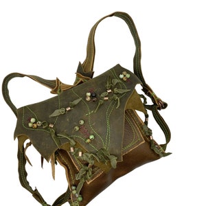 Op de afbeelding: Een groene leren rugzak met bruine leren accenten en groene en bruine kralen. De rugzak heeft een klep met een decoratieve knoop en een ritssluiting.