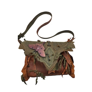 Könnte beinhalten: Handgefertigte Leder-Umhängetasche mit einem einzigartigen, von der Natur inspirierten Design. Die Tasche hat eine braune Basis, olivgrüne Akzente und einen dekorativen rosa Schmetterling. Der Riemen ist olivgrün, und die Tasche hat mehrere Schichten und Texturen.