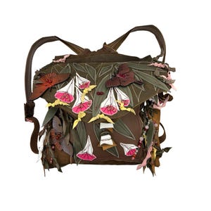 Peut inclure: Un sac à dos en cuir marron orné d'applications en cuir représentant des papillons de nuit et des fleurs blanches aux centres roses. Le sac comprend des détails en forme de feuilles vertes, des lanières en cuir pendantes et des bretelles fines.