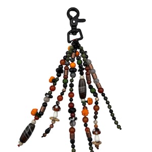 May include: A beaded keychain featuring a black lobster clasp with multiple dangling strands of beads in shades of green, orange, brown, and black. The strands include various shapes such as round beads, cylindrical beads, and small mushroom charms.