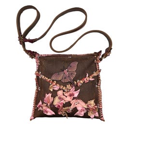 Könnte beinhalten: Braune Umhängetasche mit langem Riemen. Die Tasche hat ein Schmetterlingsdesign und rosa Schleifenakzente. Die Tasche ist quadratisch und hat eine dekorative Bordüre an den Kanten.