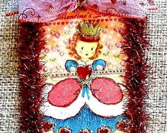 May include: A Valentine's Day ornament featuring a vintage illustration of a princess with a gold crown and a heart-shaped dress. The ornament is trimmed with red tinsel and topped with a pink ribbon bow with heart accents.