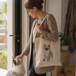 Puede incluir: Un bolso de tela beige con una ilustraci&oacute;n bordada de un gato calic&oacute;. El gato aparece sentado con patrones de pelaje marr&oacute;n, blanco y negro. El bolso tiene dos correas para el hombro y est&aacute; confeccionado con una tela de color claro.