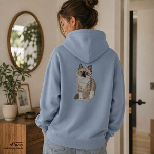 Puede incluir: Una sudadera con capucha de color azul claro que presenta un bordado de un gato esponjoso en la parte posterior. El gato tiene pelaje marr&oacute;n, tostado y blanco con marcas oscuras en la cara y las orejas. La sudadera tiene mangas largas y una capucha.