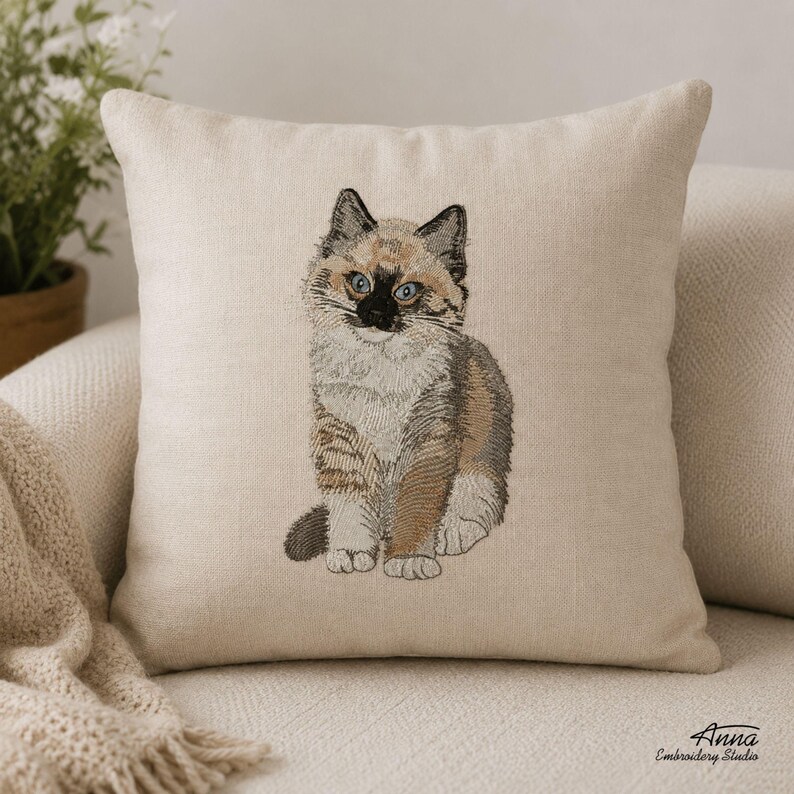 Puede incluir: Un coj&iacute;n cuadrado de color beige con una ilustraci&oacute;n bordada detallada de un gato sentado con ojos azules, pelaje marr&oacute;n y crema, y una m&aacute;scara facial oscura. En la esquina inferior derecha aparece el texto Anna Embroidery Studio en letra cursiva negra.