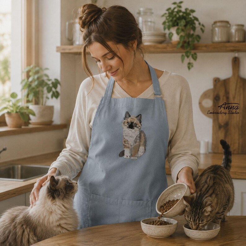 Puede incluir: Un delantal azul claro con un gato calic&oacute; bordado en el pecho. El delantal tiene una cinta para el cuello y un bolsillo frontal. El dise&ntilde;o est&aacute; centrado en la tela y muestra una representaci&oacute;n detallada de un gato con pelaje tricolor.