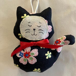 May include: A handmade black cat-shaped ornament with a white face, closed eyes, and a red bow. The cat's body is decorated with colorful floral patterns. A white string is attached for hanging.