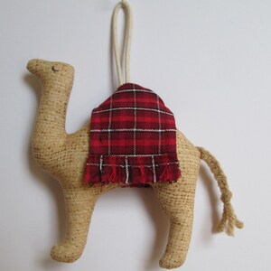 May include: A tan fabric camel ornament with a red and black plaid blanket on its back. The camel has a long, thin tail and a string loop for hanging.