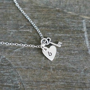 May include: A silver necklace featuring a heart pendant with the letter "b" engraved on it, accompanied by a small key charm. The necklace is displayed on a wooden surface.