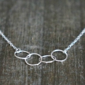 May include: A silver-toned necklace featuring a delicate chain and a central pendant composed of four interconnected circular rings. The metal has a polished finish, and the rings are linked together in a horizontal row, suspended from the thin chain.
