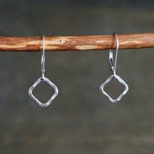 May include: A pair of silver-toned earrings. Each earring features a hammered, square-shaped design with a lever-back closure. The earrings are displayed against a blurred, neutral background, highlighting their simple elegance.