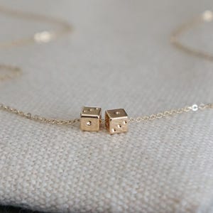 May include: A gold necklace with two small gold dice charms. The necklace is on a white fabric background.