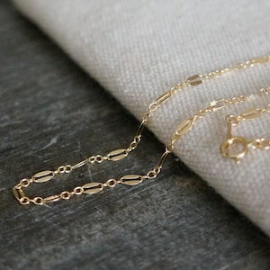 May include: A delicate gold-colored necklace with an oval link chain design. The necklace is displayed on a neutral-colored fabric, with a dark gray wooden surface in the background. The necklace has a small clasp.